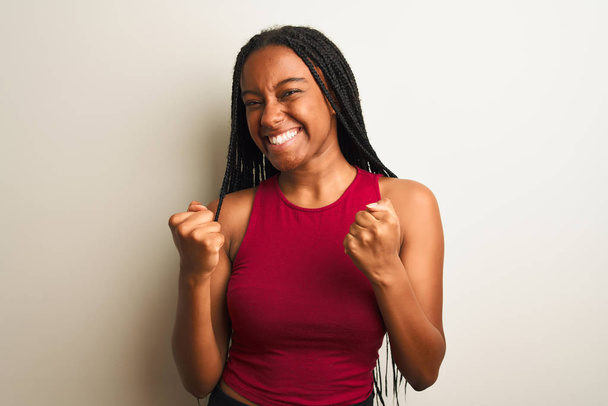 Африканська американська жінка, одягнена в червону випадкову футболку, стоїть над ізольованим білим фоном дуже щаслива і захоплена жестом переможця з піднятими руками, посміхаючись і кричачи про успіх. Святкування.. - Фото, зображення