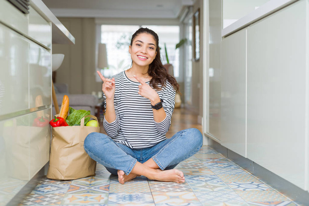 Молодая женщина, сидящая на полу кухни с бумажным пакетом, полным свежих продуктов, улыбаясь и глядя на камеру, указывая двумя руками и пальцами в сторону
. - Фото, изображение