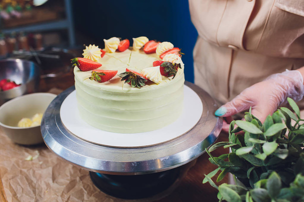 Confectioner decorated with berries a biscuit cake with white cream. Cake stands on a wooden table. The concept of homemade pastry, cooking cakes. - 写真・画像