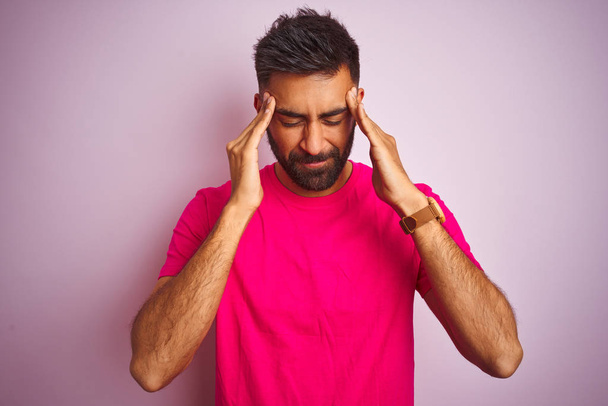 Young indian man wearing t-shirt standing over isolated pink background with hand on head for pain in head because stress. Suffering migraine. - Photo, image