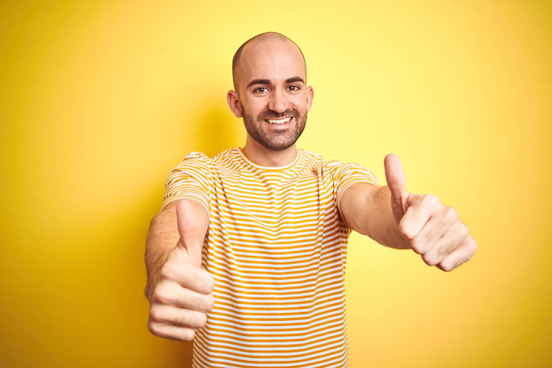 Young bald man with beard wearing casual striped t-shirt over yellow isolated background approving doing positive gesture with hand, thumbs up smiling and happy for success. Winner gesture. - 写真・画像