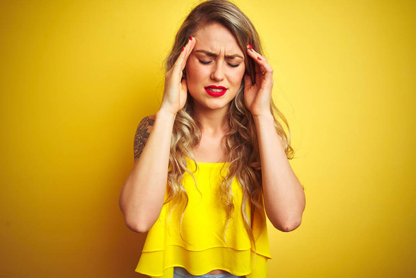 ストレスのため頭の痛みに頭を抱えて黄色の孤立した背景の上に立つTシャツを着た若い気取った女性。苦しむ片頭痛. - 写真・画像