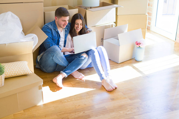 Jeune couple assis sur le sol de la nouvelle maison autour des boîtes en carton à l'aide d'un ordinateur portable, souriant heureux pour le nouvel appartement
 - Photo, image