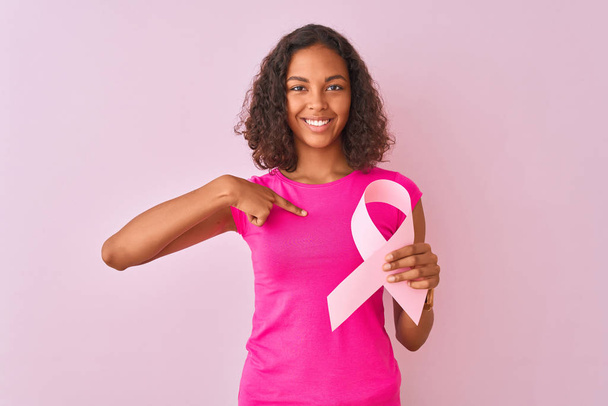 Молода Бразильська жінка тримає рак стрічку стоячи над ізольованою рожевою фон з сюрпризом особи вказуючи пальцем до себе - Фото, зображення