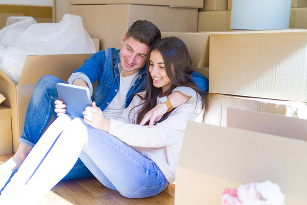 Mladí manželé sedí na podlaze nového domu kolem lepenkových krabic s tabletem, s úsměvem pro nový byt - Fotografie, Obrázek