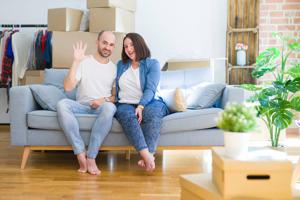 Young couple sitting on the sofa arround cardboard boxes moving to a new house showing and pointing up with fingers number five while smiling confident and happy. - Photo, Image