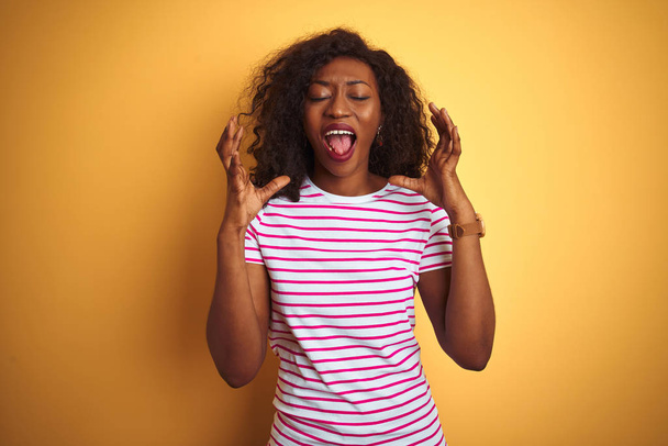 Młoda African American kobieta ubrana w paski t-shirt na białym tle żółte obchody szalony i szalony na sukces z ramion podniesione i zamknięte oczy krzyczeć podekscytowany. Koncepcja zwycięzcy - Zdjęcie, obraz