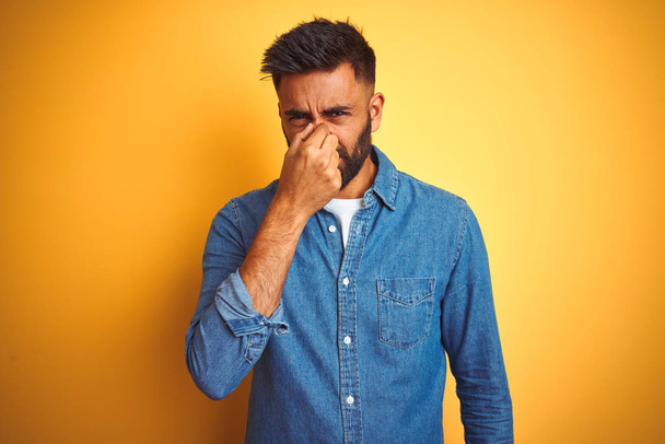 Jeune homme indien portant chemise en denim debout sur fond jaune isolé sentant quelque chose de puant et dégoûtant, odeur intolérable, retenant souffle avec les doigts sur le nez. Concept de mauvaises odeurs
. - Photo, image