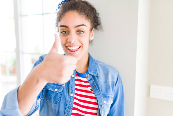 Beautiful young african american woman with afro hair wearing casual sweater doing happy thumbs up gesture with hand. Approving expression looking at the camera showing success. - Photo, Image