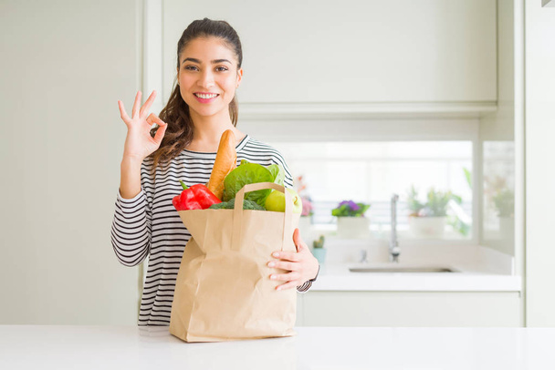 Young woman holding paper bag full of groceries doing ok sign with fingers, excellent symbol - Foto, Bild