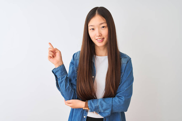 Fiatal kínai nő visel denim shirt áll át elszigetelt fehér háttér egy nagy mosollyal az arcon, mutatva a kezét, és ujját az oldalon nézi a kamerát. - Fotó, kép
