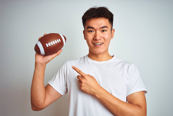 Молодой азиатский китайский спортсмен держа регби мяч стоя на изолированном белом фоне очень счастлив указывая рукой и пальцем
 - Фото, изображение