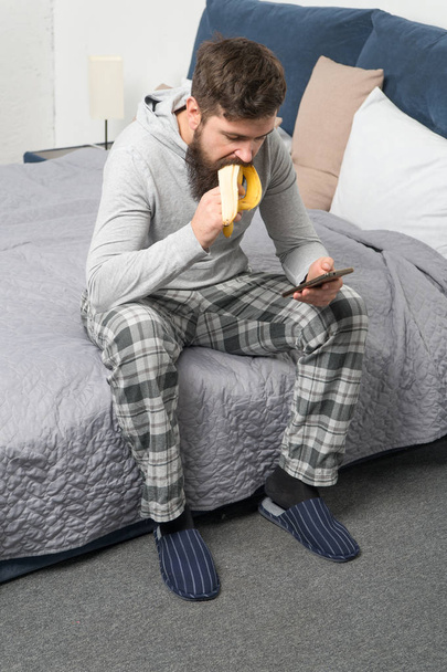 brutal sleepy man has breakfast in bedroom. mature male with beard in pajama on bed. sleep and awake. healthy lifestyle. bearded man hipster eat banana in morning. Chatting with girlfriend - Foto, Imagen