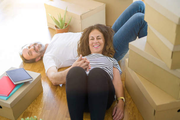 Casal romântico sênior de meia-idade deitado no chão, sorrindo feliz por se mudar para uma nova casa, relaxar e fazer uma pausa de caixas de embalagem
 - Foto, Imagem