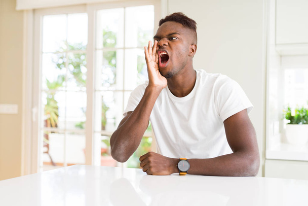 Knappe Afro-Amerikaanse man op witte tafel thuis schreeuwen en schreeuwen luid naar kant met de hand op de mond. Communicatie concept. - Foto, afbeelding