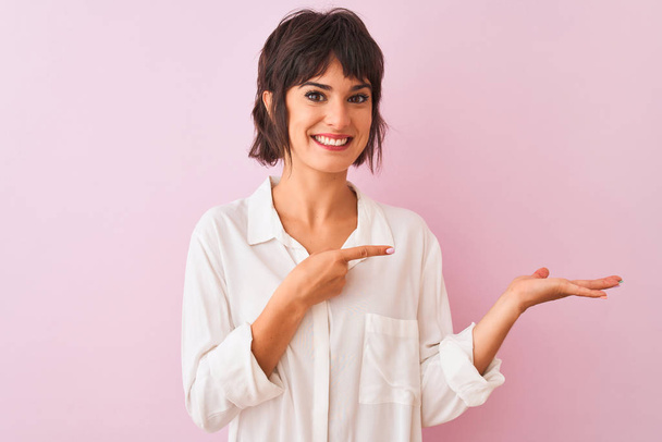 Young beautiful woman wearing white shirt standing over isolated pink background amazed and smiling to the camera while presenting with hand and pointing with finger. - Photo, Image