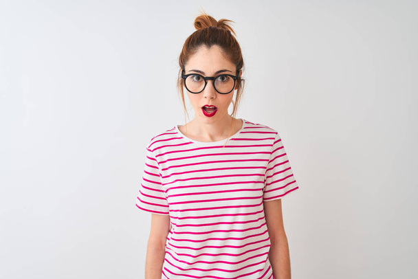 Mulher ruiva usando óculos listrados t-shirt e pigtail sobre fundo branco isolado com medo e chocado com expressão surpresa, medo e rosto animado
. - Foto, Imagem