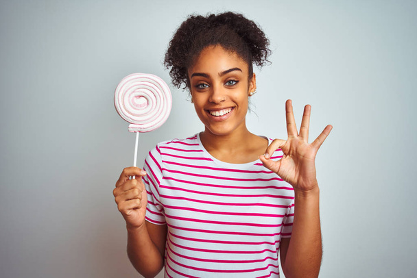 Афроамериканець підліток жінка їсть барвисті цукерки над ізольованим білим фоном робити OK підписати пальцями, відмінний символ - Фото, зображення