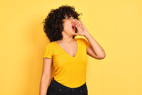junge arabische Frau mit lockigem Haar, T-Shirt vor isoliertem gelben Hintergrund stehend, schreiend und laut schreiend mit der Hand auf dem Mund. Kommunikationskonzept. - Foto, Bild
