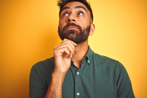 Young indian man wearing green shirt standing over isolated yellow background with hand on chin thinking about question, pensive expression. Smiling with thoughtful face. Doubt concept. - Photo, Image