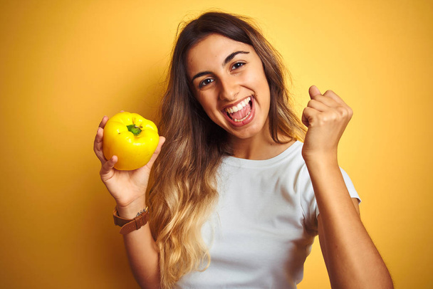 Young beautiful woman holding pepper over yellow isolated background screaming proud and celebrating victory and success very excited, cheering emotion - Фото, зображення