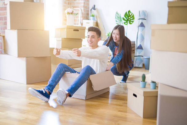 Divertida pareja asiática divirtiéndose, cabalgando dentro de una caja de cartón sonriendo feliz, muy emocionada mudándose a una nueva casa
 - Foto, Imagen