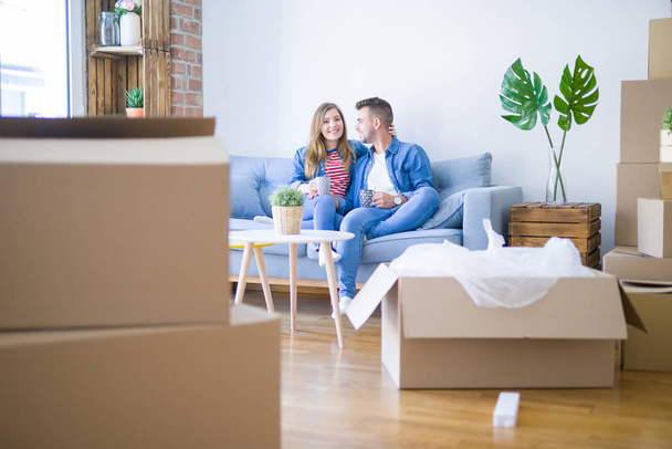 Young couple relaxing drinking a cup of coffee sitting at sofa of  new home with cardboard boxes around them, very happy moving to a new apartment - Photo, Image