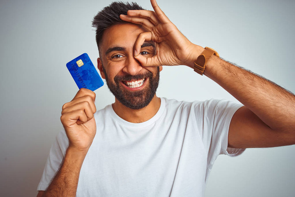 Young indian customer man holding credit card standing over isolated white background with happy face smiling doing ok sign with hand on eye looking through fingers - Photo, Image