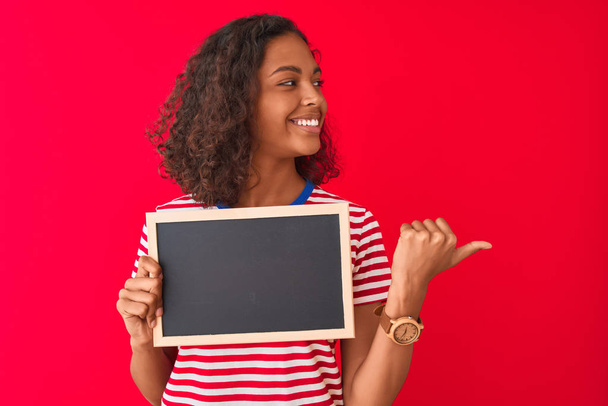 Jonge Braziliaanse student vrouw Holding Blackboard staande over geïsoleerde rode achtergrond aanwijzen en weergeven met duim omhoog naar de zijkant met vrolijk gezicht glimlachend - Foto, afbeelding
