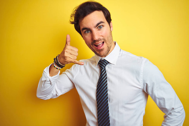 Jovem empresário bonito vestindo camisa elegante e gravata sobre fundo amarelo isolado sorrindo fazendo gesto de telefone com a mão e os dedos, como falar ao telefone. Conceitos comunicantes
. - Foto, Imagem