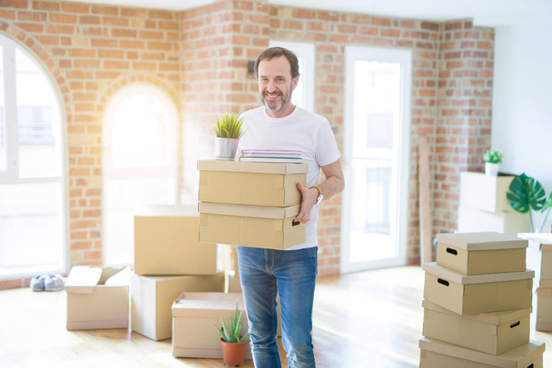 Handsome middle age senior man smiling happy moving to a new house, packing and holding cardboard boxes - Foto, Imagem