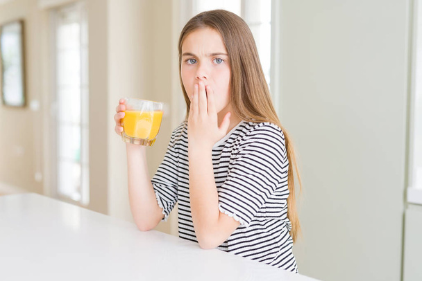 Hermosa niña bebiendo un vaso de jugo de naranja fresca cubrir la boca con la mano conmocionado con la vergüenza por error, expresión de miedo, asustado en silencio, concepto secreto
 - Foto, Imagen