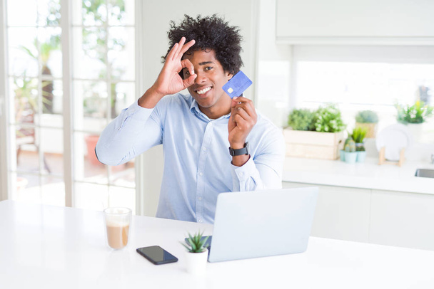 African American man using laptop and shopping online with credit card with happy face smiling doing ok sign with hand on eye looking through fingers - Fotó, kép
