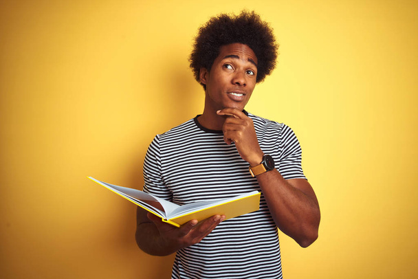 Afro american student man reading book standing over isolated yellow background serious face thinking about question, very confused idea - Foto, imagen