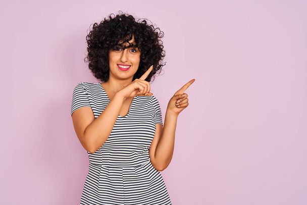 Nuori arabi nainen kihara tukka yllään raidallinen mekko yli eristetty vaaleanpunainen tausta hymyillen ja katsoen kameraa osoittaa kahdella kädellä ja sormella sivulle
. - Valokuva, kuva