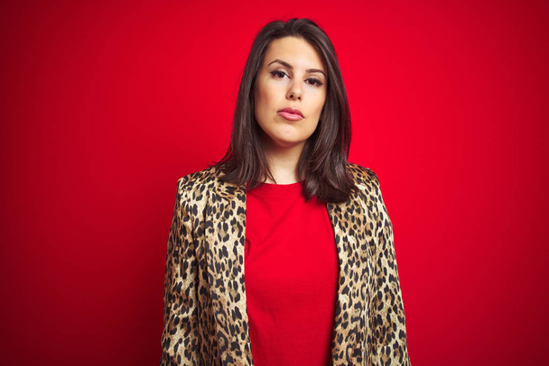 Young beautiful brunette woman wearing a leopard jacket over red isolated background with serious expression on face. Simple and natural looking at the camera. - Photo, Image