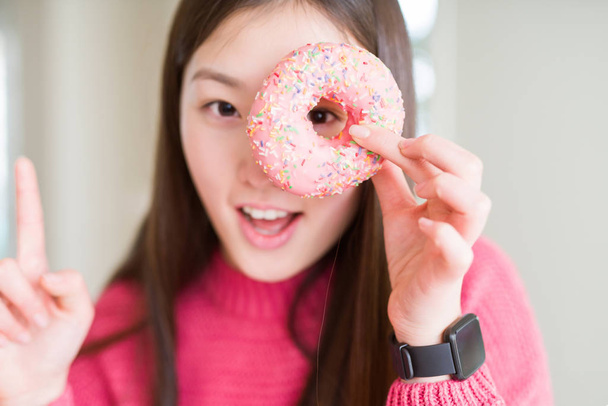 Krásná asijská žena pojídač růžového cukru překvapena myšlenkou nebo otázkou ukazující prstem se spokojeným obličejem, číslo jedna - Fotografie, Obrázek