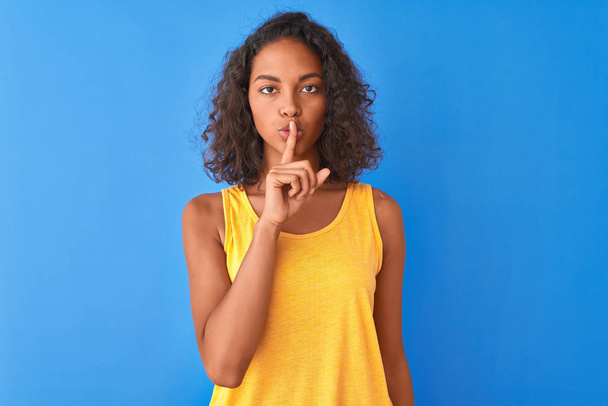 Fiatal brazil nő visel sárga póló áll az elszigetelt kék háttér kérve, hogy csendes ujjal az ajkak. Csendet és titkos koncepciót. - Fotó, kép