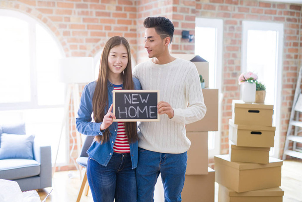 Молодая красивая азиатская пара улыбается счастливо держа доску с новым домашним текстом, обнимаясь в любви переезжая в новый дом
 - Фото, изображение