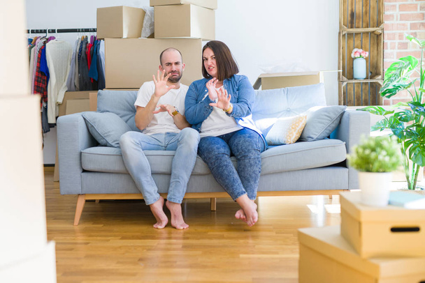 Young couple sitting on the sofa arround cardboard boxes moving to a new house disgusted expression, displeased and fearful doing disgust face because aversion reaction. With hands raised. Annoying concept. - Photo, Image