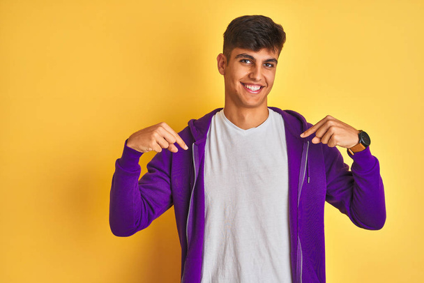 junger indischer Mann in lila Sweatshirt, der vor isoliertem gelben Hintergrund steht, sieht selbstbewusst aus, mit einem Lächeln auf dem Gesicht, zeigt mit den Fingern stolz und glücklich. - Foto, Bild