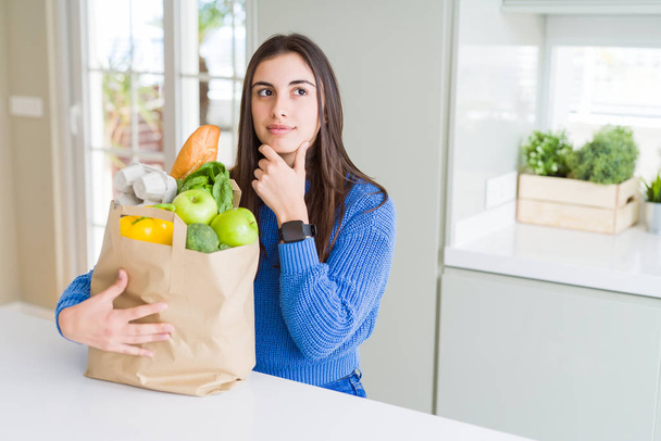 Красивая молодая женщина держит бумажный пакет, полный здоровых продуктов серьезное лицо, думая о вопросе, очень запутанная идея
 - Фото, изображение