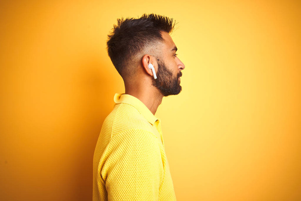 Молодий індійський чоловік слухає музику за допомогою навушників, що стоять над ізольованим жовтим тлом, дивлячись в бік, розслабляє профільну позу з природним обличчям з впевненою посмішкою
. - Фото, зображення