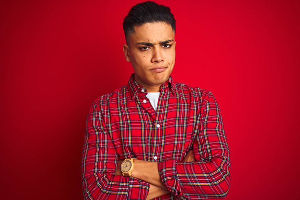 Young brazilian man wearing shirt standing over isolated red background skeptic and nervous, disapproving expression on face with crossed arms. Negative person. - Foto, imagen