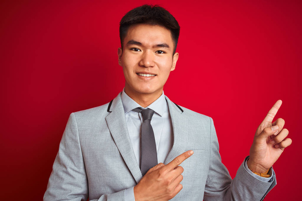 asiatischer chinesischer Geschäftsmann in grauer Jacke und Krawatte steht lächelnd vor isoliertem roten Hintergrund und blickt in die Kamera, die mit zwei Händen und Fingern zur Seite zeigt. - Foto, Bild