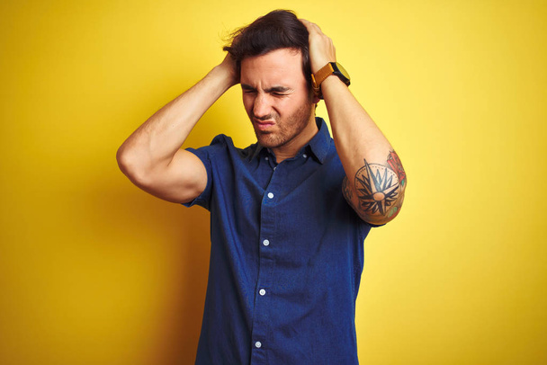 Jeune homme beau avec tatouage portant chemise décontractée debout sur fond jaune isolé souffrant de maux de tête désespérés et stressés parce que la douleur et la migraine. Mains sur la tête
. - Photo, image