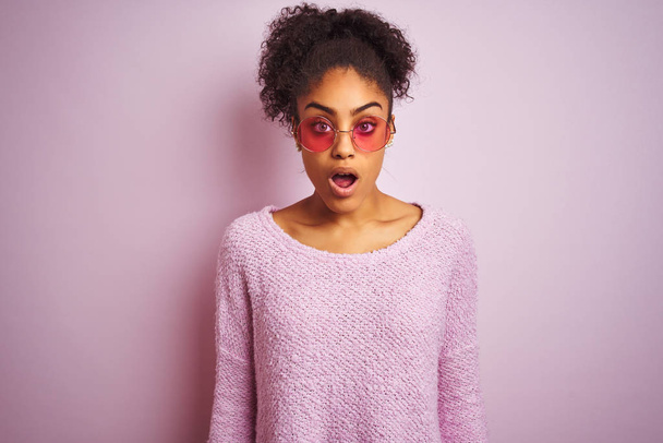 Femme afro-américaine portant un pull d'hiver et des lunettes de soleil sur fond rose isolé effrayée et choquée par l'expression surprise, la peur et le visage excité
. - Photo, image