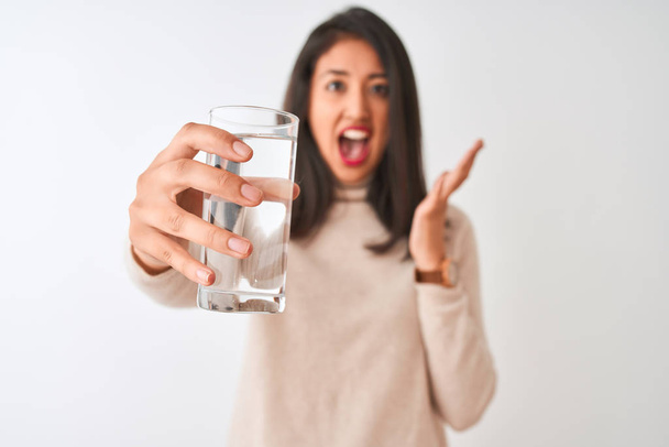 Young beautiful chinese woman holding glass of water standing over isolated white background very happy and excited, winner expression celebrating victory screaming with big smile and raised hands - Photo, Image