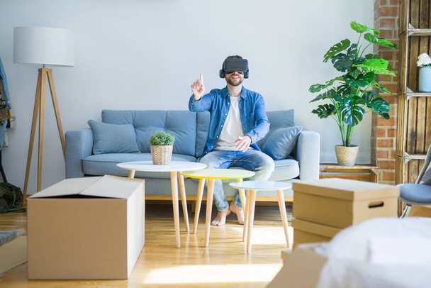 Jovem vestindo óculos de realidade virtual jogando um jogo de simulação sentado no sofá em torno de caixas de papelão se movendo para uma nova casa
 - Foto, Imagem