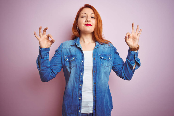 Youg beautiful redhead woman wearing denim shirt standing over isolated pink background relax and smiling with eyes closed doing meditation gesture with fingers. Yoga concept. - Photo, Image